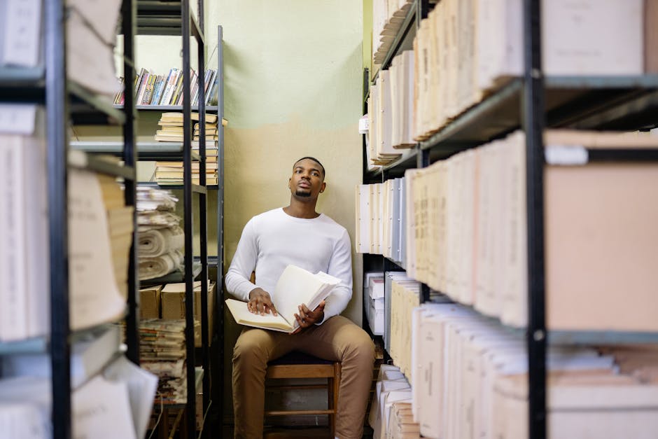 Young man sitting and reading a book in a library archive room.
